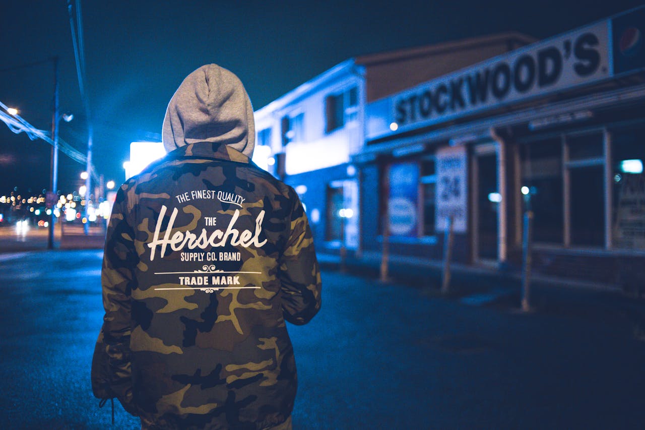 Hooded man in camo jacket stands on an urban street at night, captured in moody lighting.